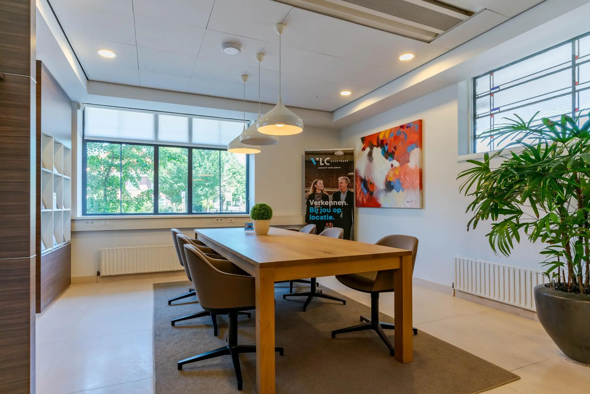 Moderne vergaderruimte aan de Van Alkemadelaan met houten tafel, designstoelen, kunst aan de muur en grote ramen met uitzicht op groen.